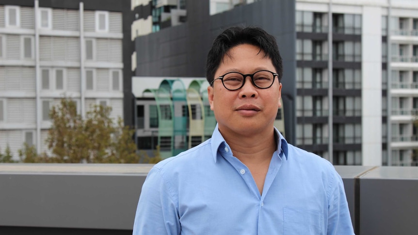 Robertus Robet wears a blue shirt and stands in front of office towers while looking at the camera.