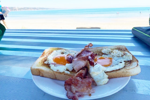 A plate of bacon and eggs with toast, on a table overlooking the beach in the background 
