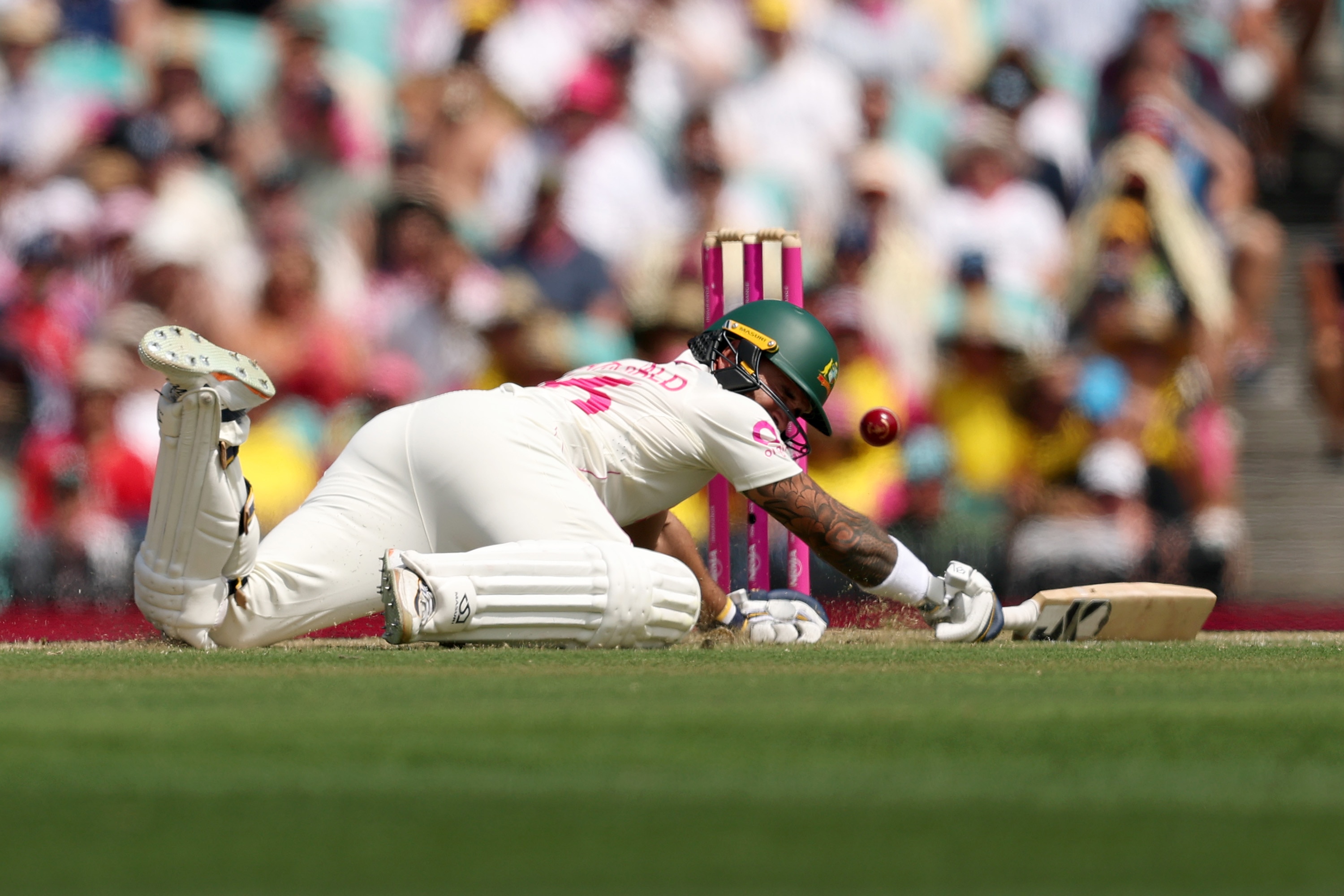 Um batedor de críquete de branco mergulha em sua área com a bola voando em direção aos tocos