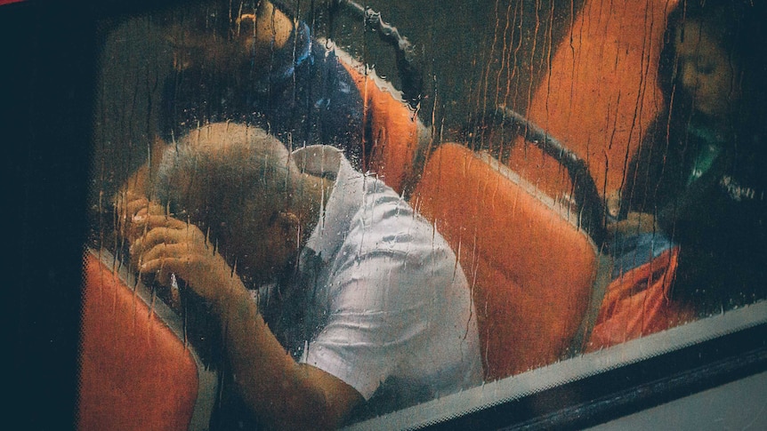 A man is seen through a rain-streaked bus window with his head in his hands on the seat in front of him