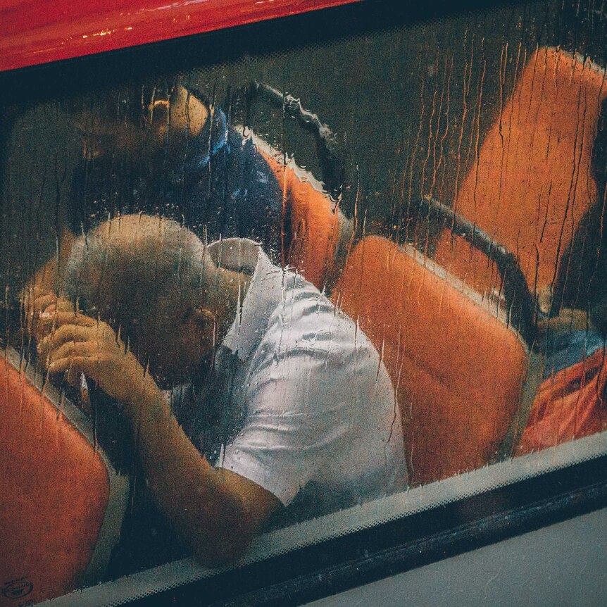 A man is seen through a rain-streaked bus window with his head in his hands on the seat in front of him