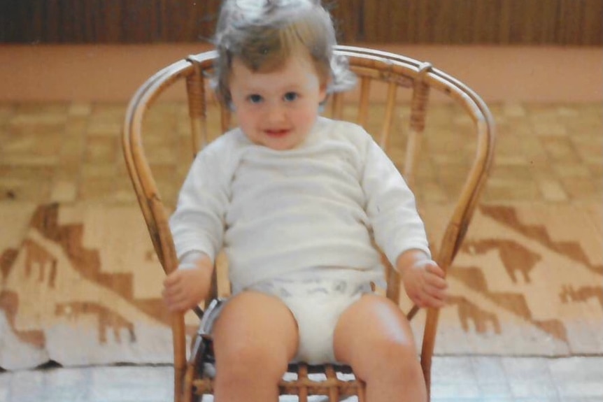 Kate Drysdale as a toddler, sitting in a cane chair, wearing white.