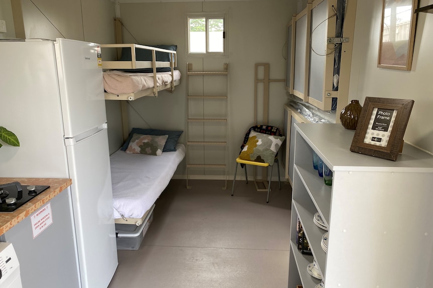 A narrow room showing the edge of a kitchen stove, a fridge, two bunk beds and some collapsible benches.