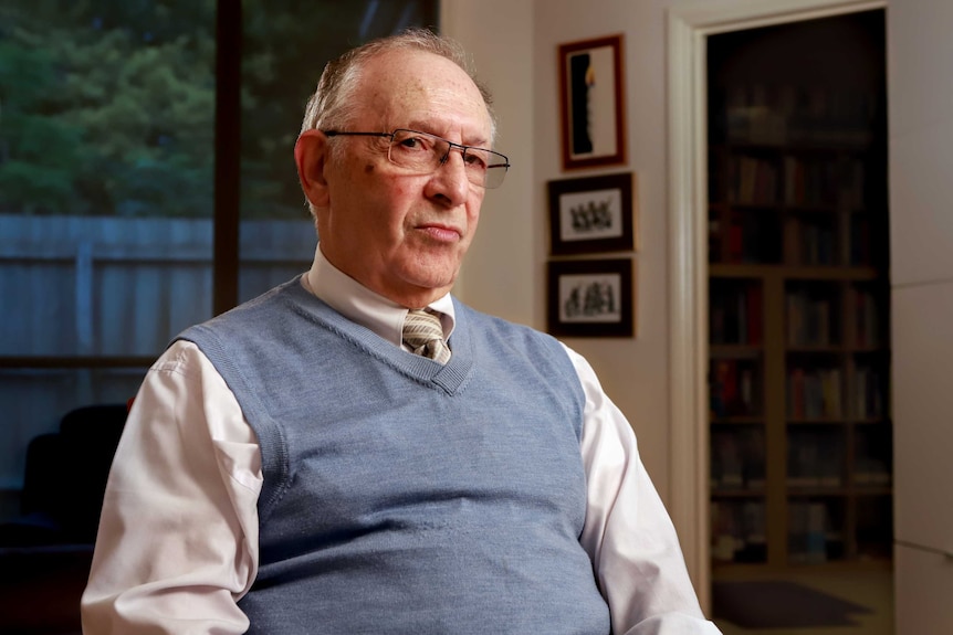 An older man in a white shirt and blue vest,points at his computer in his study.