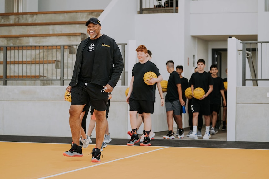 Tracy Williams smiles as he leads a group of boys holding basketballs onto an indoor court.