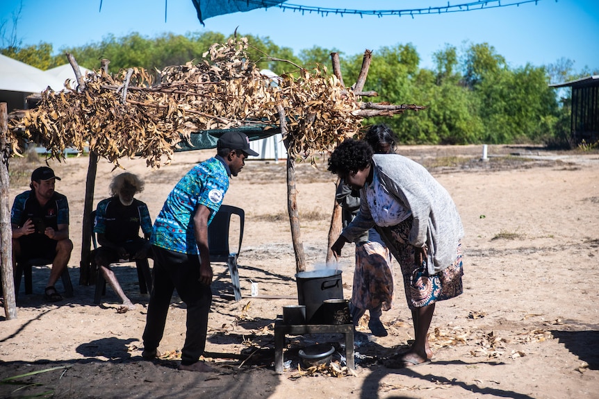 People surround a pot on a camp fire under a blue sky and sand, trees in the distance.