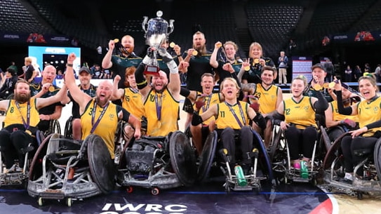 The Australian wheelchair rugby team gather on court for a celebratory group shot, holding the trophy after a tournament win.