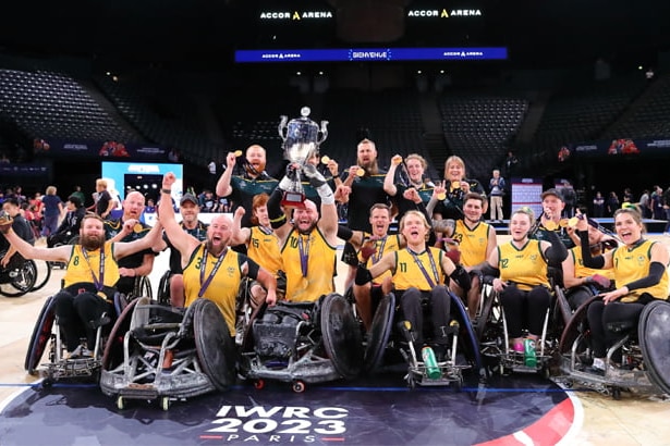 The Australian wheelchair rugby team gather on court for a celebratory group shot, holding the trophy after a tournament win.