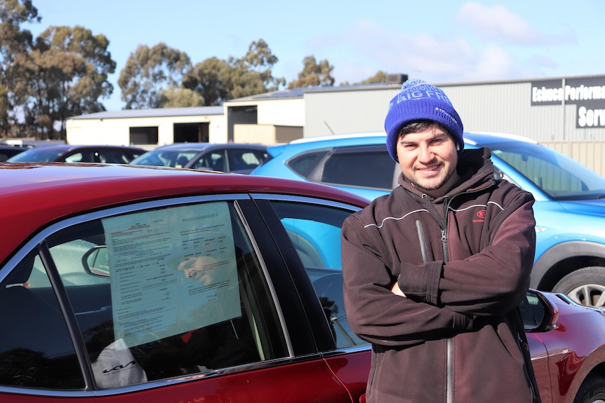 Man stands against red car with arms crossed and smiling at camera.