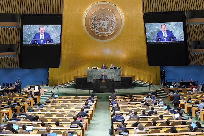 A man in a suit addresses a hall of people against a gold backdrop with two screens on either side