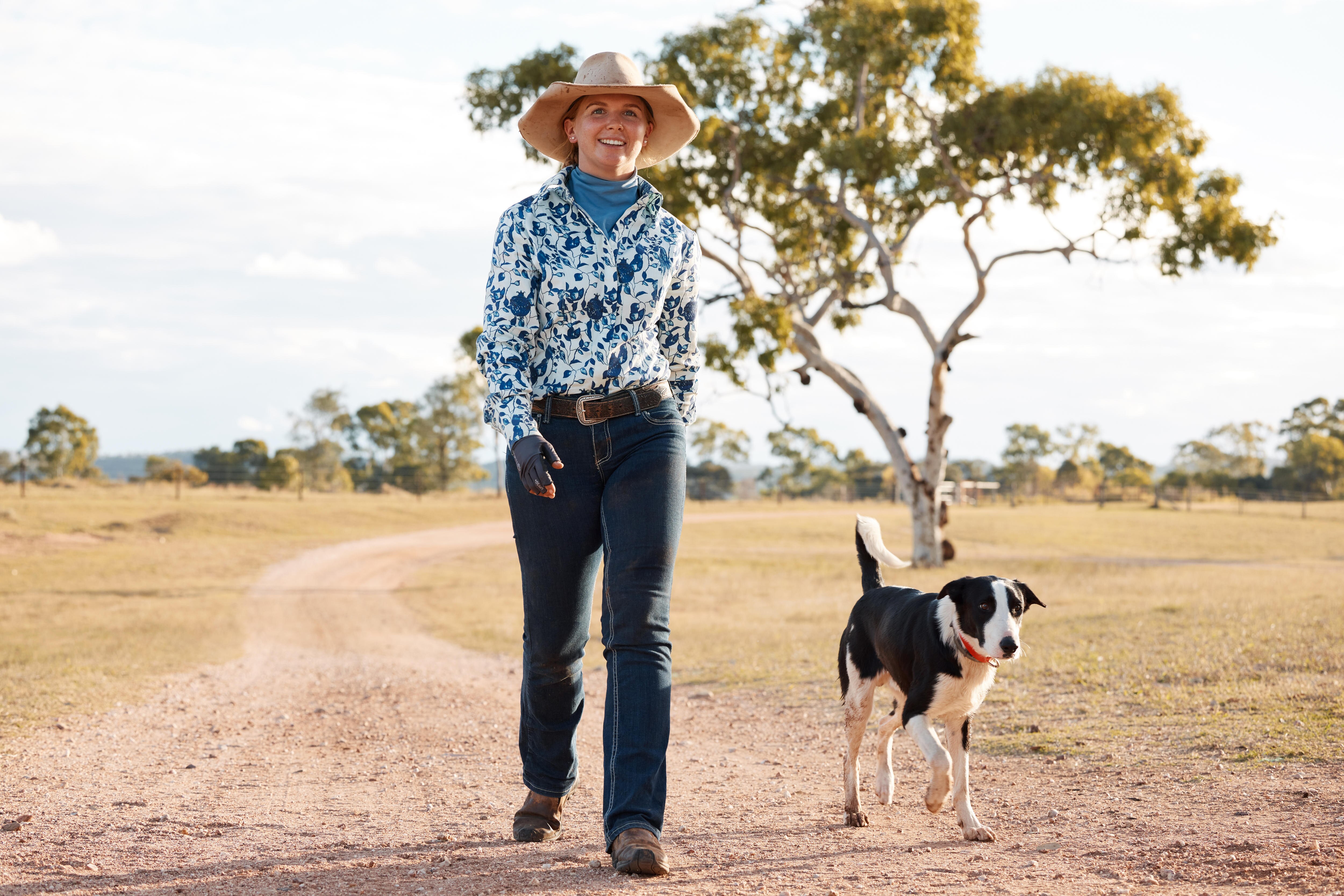 A smiling woman in farm clothes strides down a path with her dog.