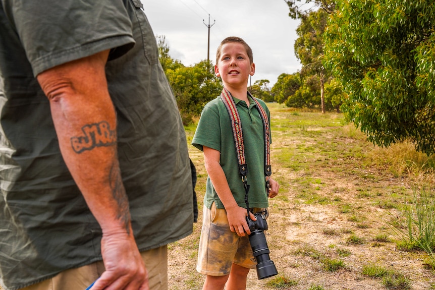 Archer Leddy turns to his Dad, Luke, while birding. Dad, partially visible, has a tattoo on his arm. Archer is carrying a camera