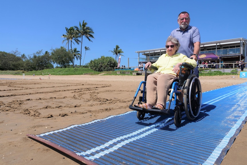 An elderly woman in a wheelchair being pushed along blue beach matting by her carer. 