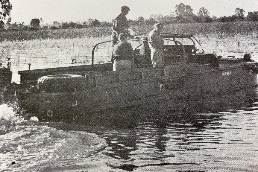 Three men ride in the back of a vehicle driving through water