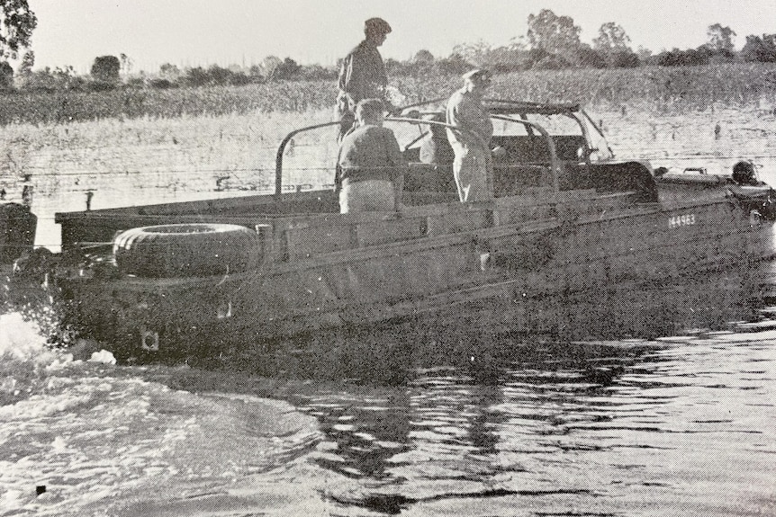 Three men ride in the back of a vehicle driving through water