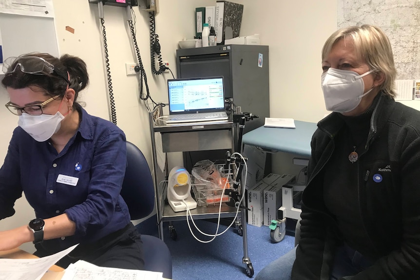 A woman in her sixties in a mask beside a doctor typing on computer