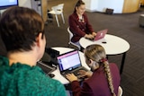 Two female high school students use a laptop at desks with a teacher overseeing.
