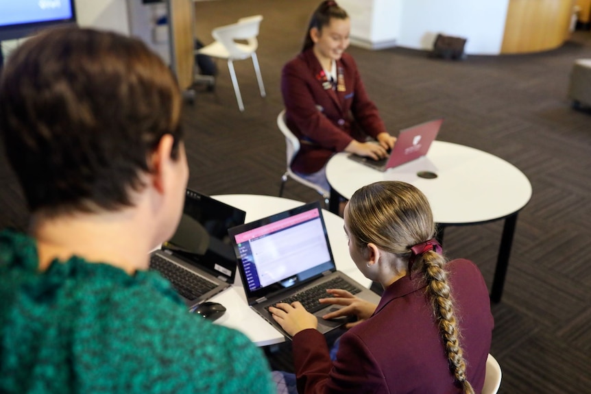 Two female high school students use a laptop at desks with a teacher overseeing.