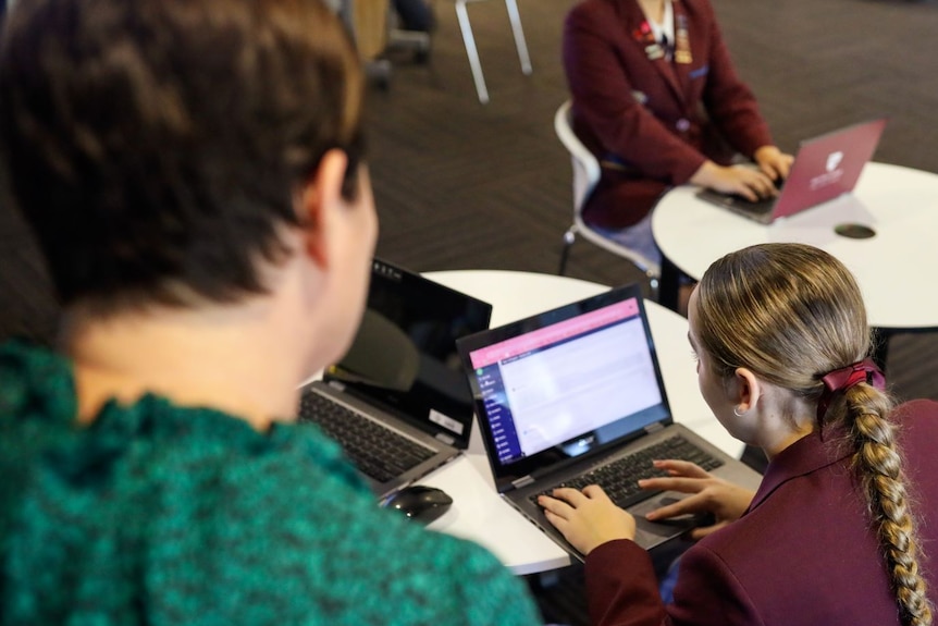 Two female high school students use a laptop at desks with a teacher overseeing.