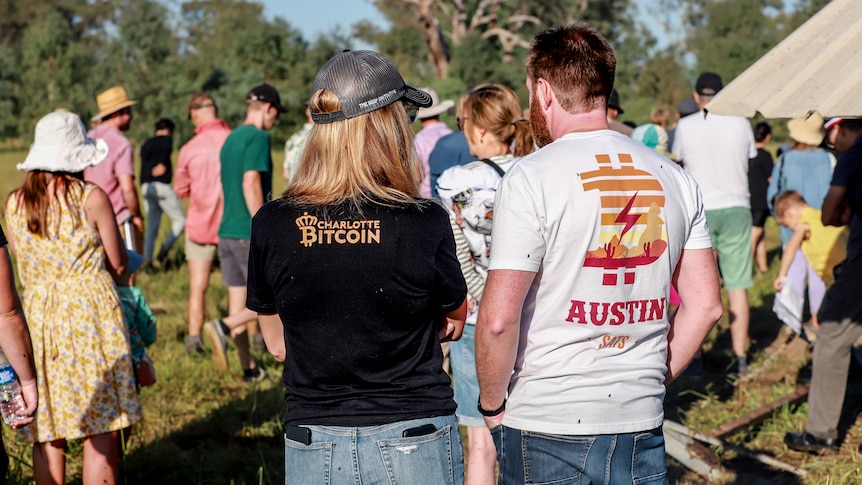 Two people stand in foreground with Bitcoin themed tshirts, while a group of several dozen mill about at a farm