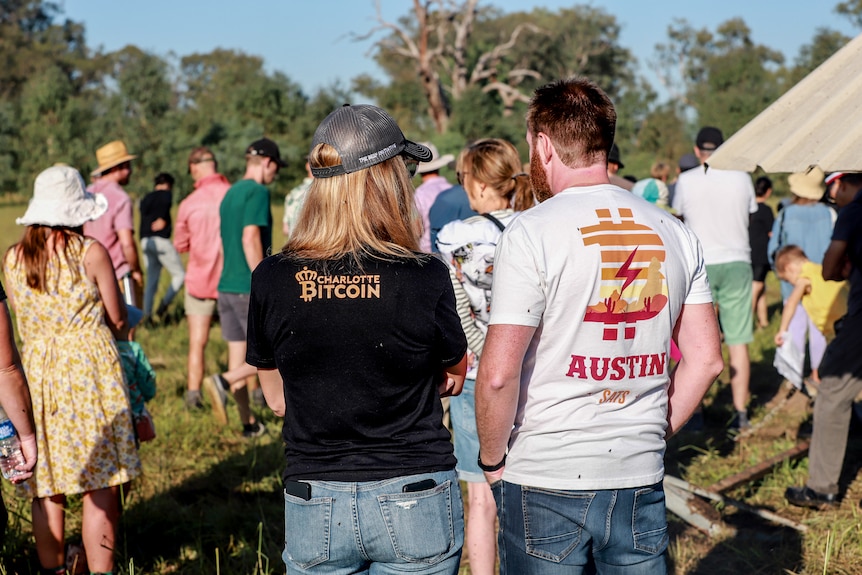 Two people stand in foreground with Bitcoin themed tshirts, while a group of several dozen mill about at a farm
