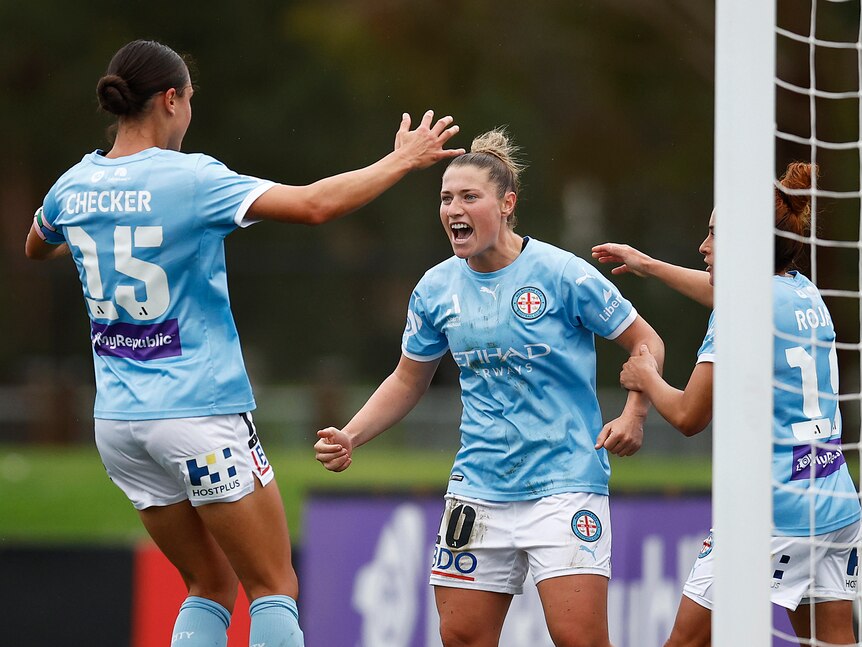 A soccer player wearing light blue and white yells as her team-mates celebrate after a goal