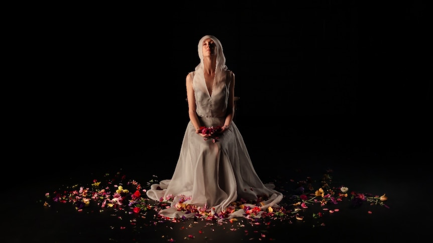 A woman sits holding flowers wearing a silver organza dress with a veil. Flowers are strewn about her feet.