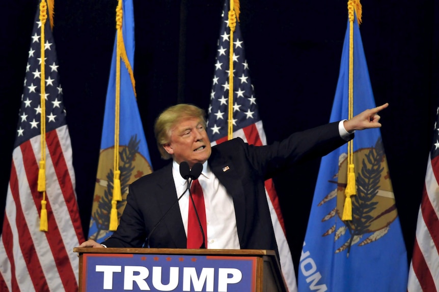 Donald Trump standing in front of flags points off to the side.