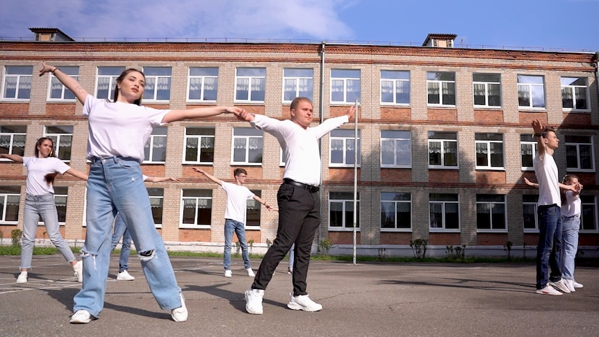 Boys and girls in pairs hold their arms out in a flourish as they dance on asphalt by a school building