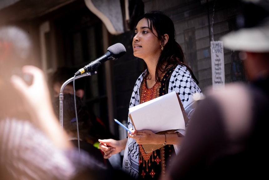 A girl with dark hair, wearing a Keffiyeh, holding a notebook and pen, speaks into a microphone on stage.