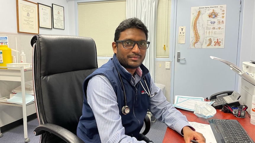 A doctor sits at his desk with a picture of a spine on the door behind him