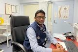 A doctor sits at his desk with a picture of a spine on the door behind him