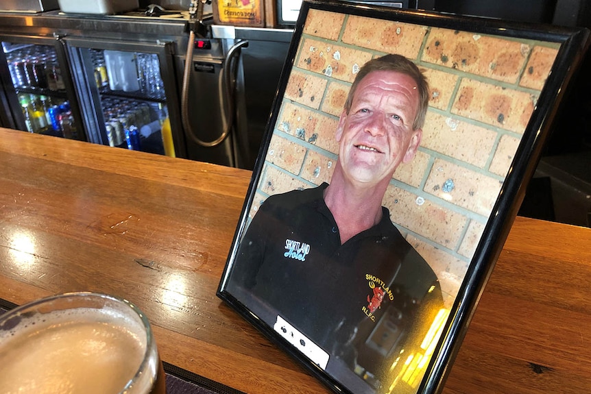 A framed photograph of a man in his 50s, sitting on the bar of a pub.