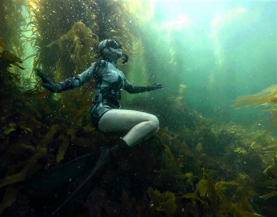 A woman free diving, with her legs curled up, looking towards the surface