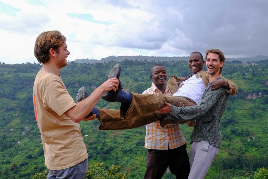 Three men holding up another man against a backdrop of rolling lush green hills.