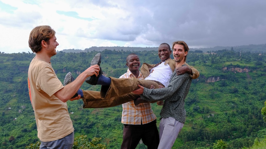 Three men holding up another man against a backdrop of rolling lush green hills