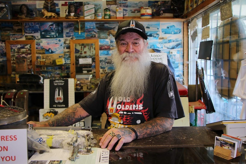 A man with a long grey beard wearing a Holden cap and shirt surrounded by Holden memorabilia