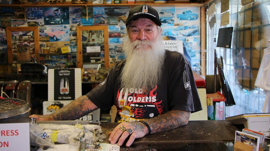 A man with a long grey beard wearing a Holden cap and shirt surrounded by Holden memorabilia