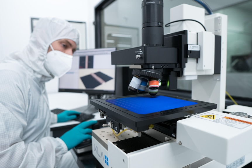 Un homme vêtu d'une combinaison de protection blanche travaille sur une cellule solaire bleue à l'intérieur d'un laboratoire.