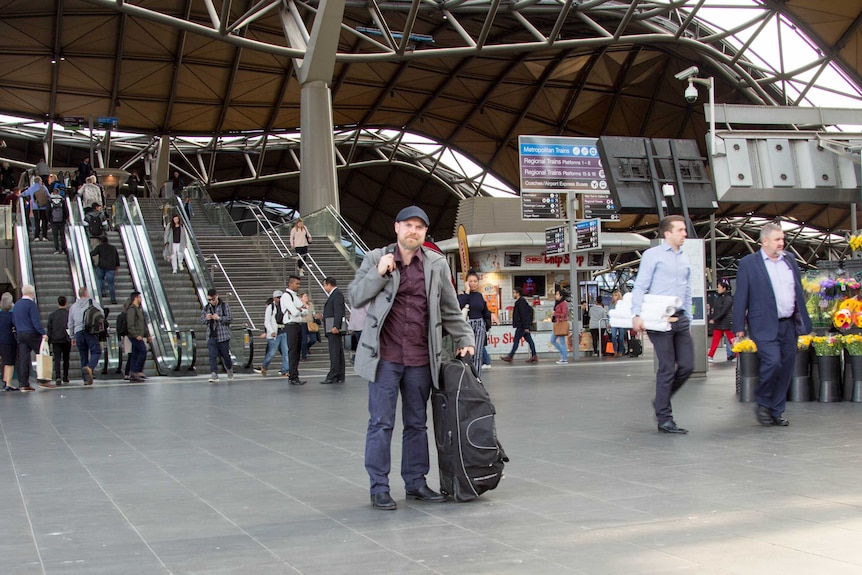 A man stands with baggage on the concourse of a large railway station.