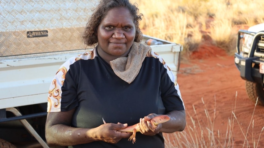 Aboriginal woman Janice Carrol holds a red and gold lizard in front of a ute in the desert.