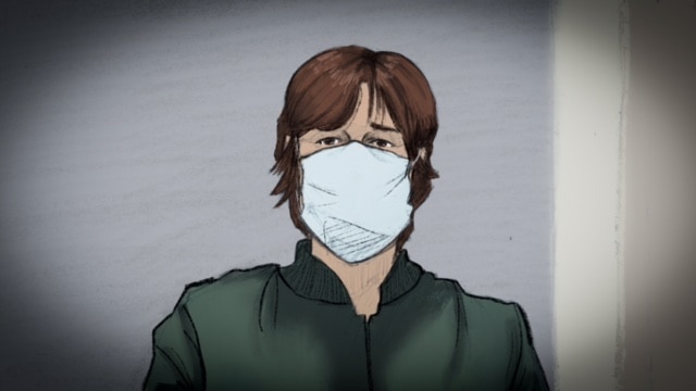 A man with longish brown hair sits against a grey wall, dressed in green and wearing a surgical mask.