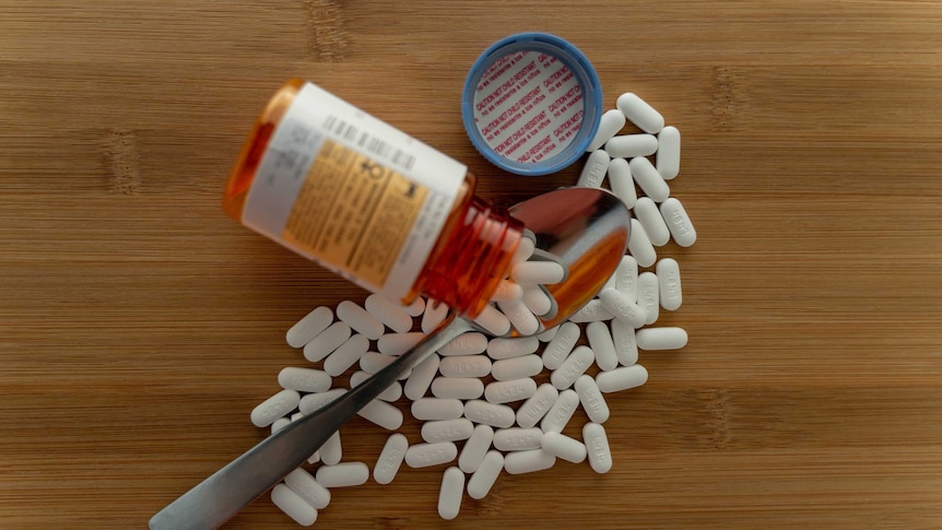Dozens of white capsules spill out of a medical bottle onto a wooden table.