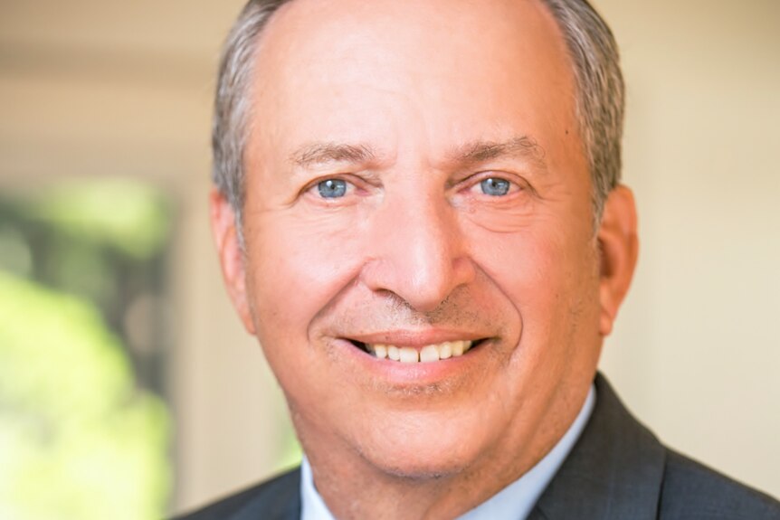 Larry Summers, a middle aged man dressed in a dark suit and blue shirt, poses for a photo smiling