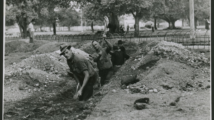black and white image of two men diggining a trench with fence and trees in background