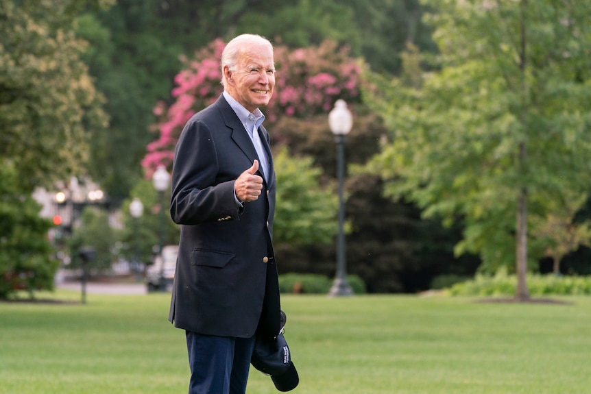 US President Joe Biden wearing a suit, smiling and giving a thumbs up while walking on grass
