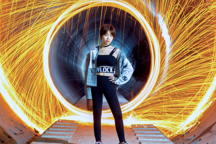 A young Chinese woman standing in a tunnel with sparks behind her.