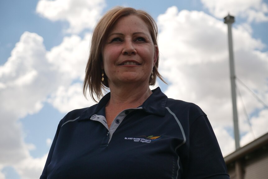 A woman with short blonde hair wearing a Bland Shire Council polo shirt looks into the distance with clouds behind her.