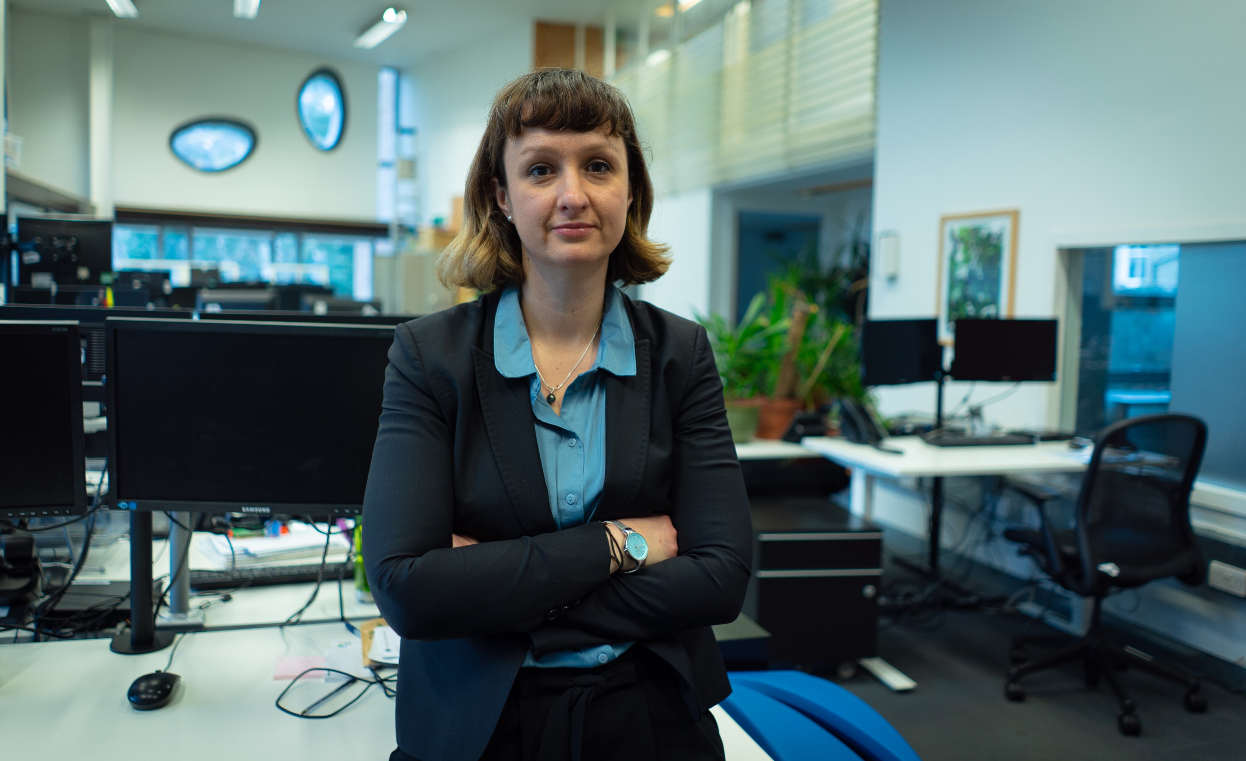 Woman wearing a dark jacket and blue shirt with her arms folded standing in an office.