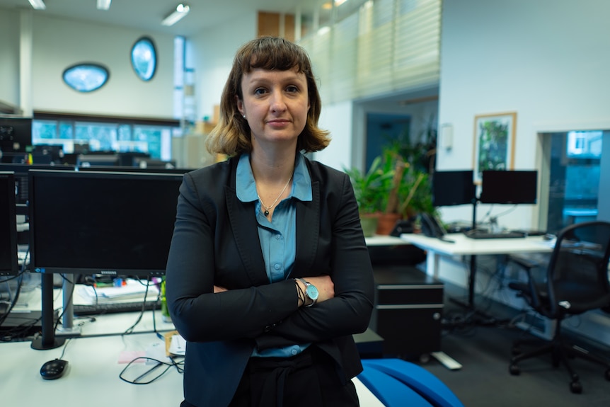 Woman wearing a dark jacket and blue shirt with her arms folded standing in an office.